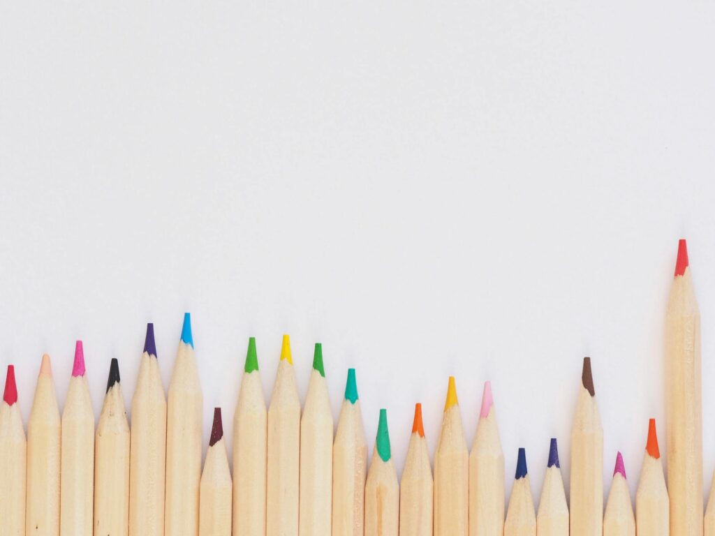 A row of colored pencils with natural wood casings aligned vertically on a white background. Each pencil has a different colored tip, creating a vibrant spectrum. The arrangement varies in height, adding a dynamic effect.